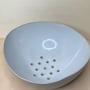 May include: A light blue ceramic colander with a wide, shallow bowl shape. The colander has a series of small holes at the bottom for draining and a slightly raised rim. The bowl is approximately 12 inches in diameter.