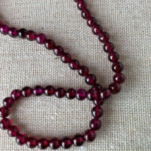 May include: A strand of deep red, translucent, spherical garnet beads arranged in a loose loop. The beads are uniform in size and shape, featuring a polished, reflective surface that highlights the rich, dark crimson colour of the natural gemstone.