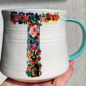 May include: A white ceramic mug with a light blue interior and handle, featuring a large letter T formed by colorful floral patterns, green vines, and a brown rabbit with a red ribbon. The mug has a ribbed texture along the exterior surface.