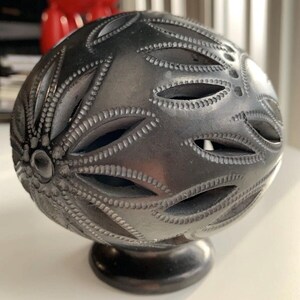 May include: A dark grey, spherical decorative object with intricate, cut-out floral patterns. The object sits on a small, round base. The design features detailed lines and textures, creating a unique, artistic appearance.