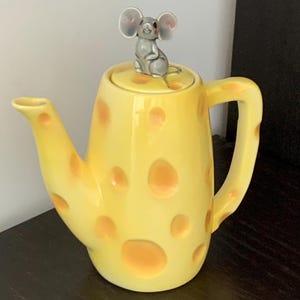 Puede incluir: Una tetera de cerámica amarilla diseñada para parecer un bloque de queso suizo con agujeros por todo el cuerpo, el asa y el pico. La tapa presenta una pequeña figura de ratón gris esculpida en la parte superior, que sirve como asa.