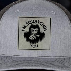 May include: Grey trucker cap with a black mesh back panel and a square patch. The patch displays a black Bigfoot illustration pointing, with the text "I'M SQUATCHIN YOU". The cap has a curved peak and a black leatherette front.