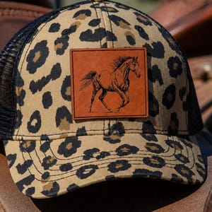 May include: A baseball cap featuring a tan and black leopard print pattern with a black mesh back. A square brown leatherette patch is centred on the front, displaying a detailed black line drawing of a horse in a running motion.