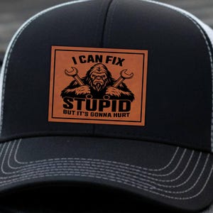 Peut inclure: Casquette de camionneur noire avec un dos en filet blanc. Le devant arbore un patch en similicuir marron avec le texte "I CAN FIX STUPID BUT IT'S GONNA HURT" et un dessin de Bigfoot tenant des clés. Accessoire humoristique.