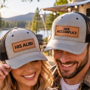 May include: Two heather gray and black mesh trucker hats featuring rectangular tan leatherette patches. One hat displays the text HIS ALIBI in black, while the other hat displays the text HER ACCOMPLICE in black. Both hats have a structured front.