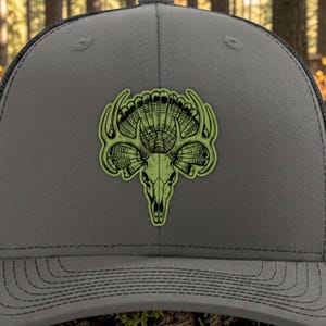 Peut inclure: Une casquette de baseball gris anthracite avec un dos en maille noire et un écusson en similicuir vert sur le devant. L'écusson présente une illustration graphique d'un crâne de cerf intégré à une queue de dinde. Le texte indique green/black leatherette charcoal/black mesh hat.