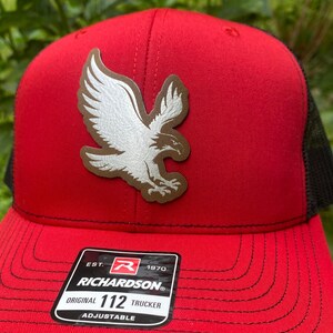 May include: A red trucker hat featuring a black mesh back and a brown leatherette patch on the front. The patch displays a white eagle in flight. A black sticker on the brim reads EST. 1970 RICHARDSON ORIGINAL 112 TRUCKER ADJUSTABLE.