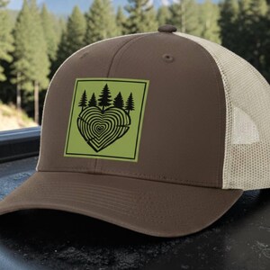 May include: A brown and khaki mesh trucker hat featuring a square green leatherette patch on the front. The patch displays a black graphic of a heart shaped like tree rings topped with a silhouette of pine trees against a solid green background.