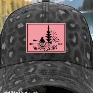 May include: A black baseball cap with a dark gray leopard print pattern and a black mesh back. The front features a rectangular pink patch with a black graphic illustration of a person kayaking on water near a tall pine tree and mountains.