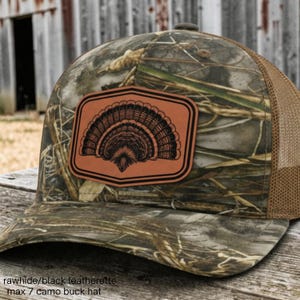 May include: A camouflage baseball cap featuring a brown leatherette patch on the front. The patch displays a black engraved graphic of a fanned-out turkey tail feather. The cap includes a brown mesh back and a curved brim with a camo pattern.