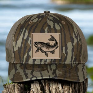 May include: A brown and green camouflage trucker hat with a brown patch featuring a black and white illustration of a fish.