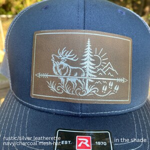 May include: A navy blue baseball cap with a rectangular brown patch featuring a laser-engraved design of an elk, a pine tree, a sun, mountains, and an arrow. The hat includes a black sticker on the brim with the letter R and the text EST 1970.