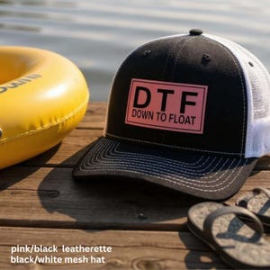 May include: Black and white mesh hat with a pink rectangular patch that reads "DTF DOWN TO FLOAT". The hat is on a wooden surface next to a yellow inflatable tube and a pair of flip-flops. The hat has a curved brim.