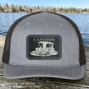 May include: A heather gray baseball cap with a black mesh back and a rectangular black leatherette patch on the front. The patch features a white graphic of a pontoon boat above the text CAPTOON, all enclosed within a rope-style border.