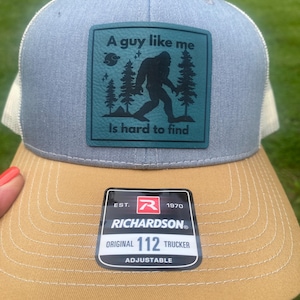 May include: A light blue and tan trucker hat featuring a rectangular teal patch with a black silhouette of Bigfoot, pine trees, and the text A guy like me is hard to find. A black sticker on the tan brim reads Richardson Est. 1970 Original 112 Trucker.