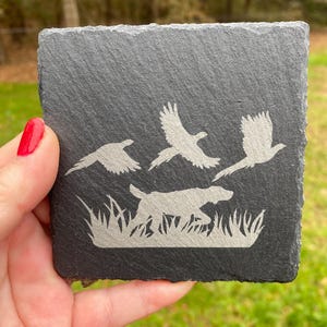May include: A black slate coaster with a white silhouette of a dog running through tall grass and chasing three birds. The birds are flying in a line.