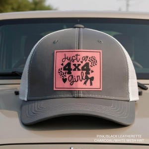 May include: Gray trucker hat with a white mesh back and a pink square patch. The patch features the text "Just a 4x4 Girl" in black script and a bow. The hat is sitting on the hood of a vehicle.