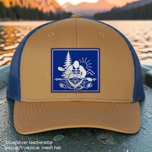 May include: A biscuit-colored baseball cap with a blue mesh back and a blue leatherette patch on the front. The patch features a white graphic of a figure kayaking with a pine tree, sun, and mountains, framed by a horizontal arrow design.