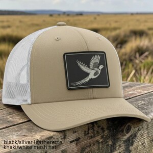 May include: A khaki and white mesh trucker hat featuring a rectangular black and silver leatherette patch on the front. The patch displays a detailed graphic of a bird in flight. The hat has a solid khaki front panel and a white mesh back.
