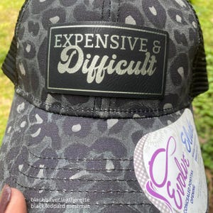 May include: Black leopard print mesh hat with the words "EXPENSIVE & Difficult" in silver lettering on a black leatherette patch. The hat has a curved brim and a concealed ponytail opening.