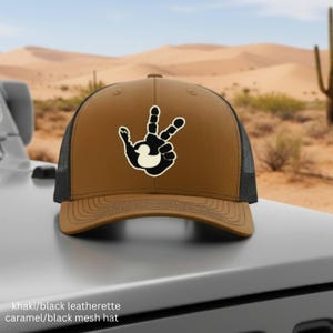 May include: Khaki and black trucker hat with a black and white hand graphic. The hat has a brown front panel and a black mesh back. The text on the image says "khaki/black leatherette caramel/black mesh hat."
