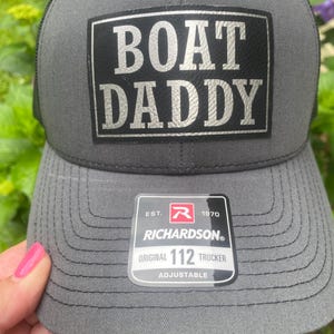 Peut inclure: Casquette de camionneur grise avec le texte "BOAT DADDY" en argenté sur un patch noir. Le chapeau a un dos en filet et une visière incurvée. L'étiquette de la marque Richardson est visible.