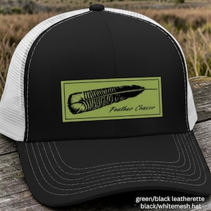 May include: A black baseball cap with a white mesh back and a black brim featuring decorative stitching. The front panel displays a rectangular olive green patch with a black feather graphic and the text Feather Chaser printed in black.