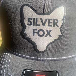 May include: A gray and black trucker hat with a silver fox logo and the text "SILVER FOX" on the front. The hat also has a red and white label with the text "EST. R 1970" on the side.