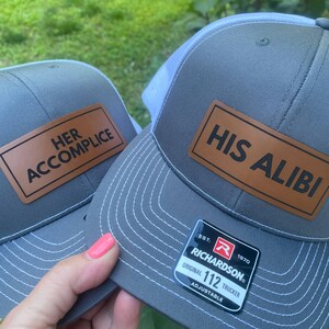 May include: Two gray trucker hats with white mesh backs. One hat has a brown leather patch that reads "HER ACCOMPLICE", the other reads "HIS ALIBI". Both hats have a Richardson tag.