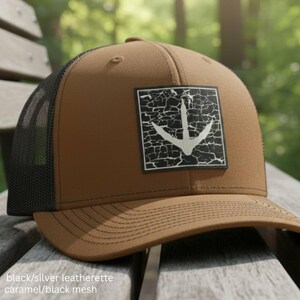 May include: A caramel-colored baseball cap featuring a black mesh back and a square patch on the front. The patch displays a white bird footprint graphic set against a black background with a distressed, cracked texture pattern.