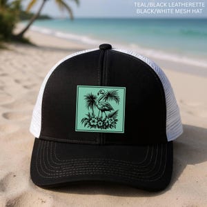May include: Black and white mesh hat with a flamingo design. The hat features a black front panel with a square teal patch showcasing a flamingo, palm trees, and flowers. The back of the hat is white mesh.