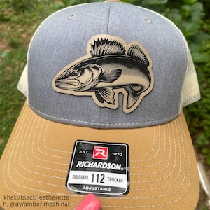 May include: Gray and amber mesh trucker hat with a fish leatherette patch. The hat features a curved brim and an adjustable snap closure. The patch is a detailed illustration of a fish. The hat has the Richardson 112 Trucker label.