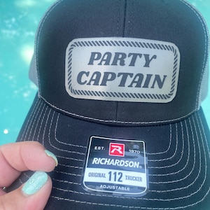 May include: Black trucker hat with a gray mesh back and a silver patch that reads "PARTY CAPTAIN." The hat has white stitching and a Richardson tag that says "ORIGINAL 112 TRUCKER."