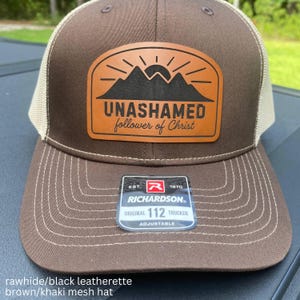 May include: A brown and khaki mesh trucker hat featuring a tan leatherette patch on the front. The patch displays a black mountain and sun graphic with the text UNASHAMED follower of Christ. A sticker on the brim reads RICHARDSON 112 TRUCKER.