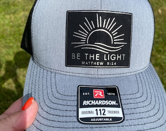 May include: A grey and black trucker hat featuring a black rectangular patch with a white sun graphic and the text BE THE LIGHT MATTHEW 5:14. A black sticker on the brim displays the text RICHARDSON ORIGINAL 112 TRUCKER EST. 1970 ADJUSTABLE.