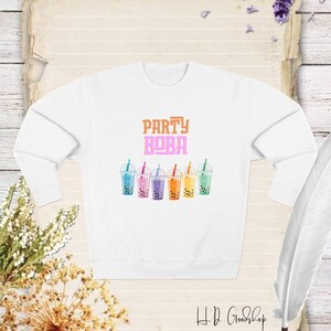 以下が含まれることがあります： オレンジとピンクの文字で「Party Boba」と書かれた白いスウェットシャツ。グラフィックには、ストローが付いたカラフルなボバティーカップが6つ描かれています。