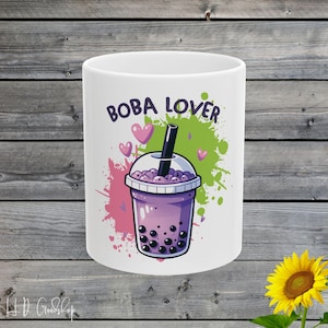 以下が含まれることがあります： ピンク、グリーン、ホワイトの飛び散ったデザインが施された白いセラミックマグカップ。マグカップには黒い文字で「BOBA LOVER」という文字と、黒いタピオカパールが入った紫色のボバティーの漫画の画像が描かれています。