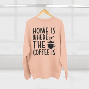 May include: A peach-colored sweatshirt with the text "Home is where the coffee is" in black lettering. A coffee cup with a mustache is above the word "the".