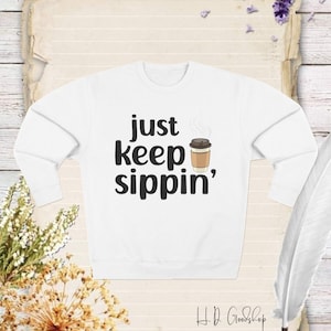 Puede incluir: Sudadera blanca con texto negro que dice "just keep sippin'" y una imagen de dibujos animados de una taza de café con vapor saliendo de ella.
