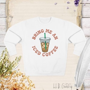 Könnte beinhalten: Weißer Pullover mit einem grafischen Design eines Bechers mit Eiskaffee und dem Text "Bring me an iced coffee" in einer Retro-Schrift.
