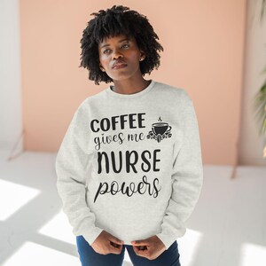 Può includere: Una felpa grigia chiaro con il testo "COFFEE gives me NURSE powers" in nero. Una piccola tazza di caffè nera con vapore è sopra la parola "NURSE".