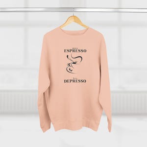 Puede incluir: Una sudadera de color melocotón con el texto "more ESPRESSO less DEPRESSO" y una imagen estilizada de una taza de café con vapor.