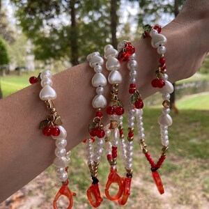 Peut inclure: Plusieurs bracelets de perles avec des perles blanches, des perles en forme de c&oelig;ur et des breloques de cerises rouges. Les bracelets ont des accents dor&eacute;s et des fermoirs rouges. L'arri&egrave;re-plan est une sc&egrave;ne ext&eacute;rieure floue avec des arbres.