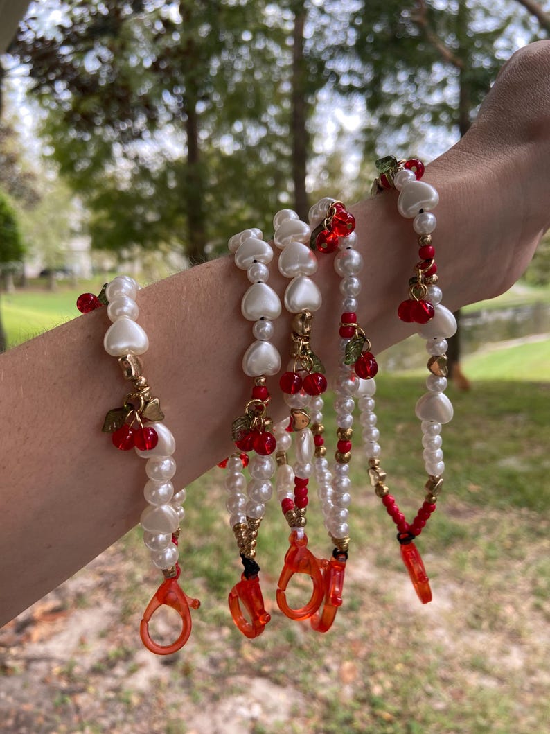 Peut inclure: Plusieurs bracelets de perles avec des perles blanches, des perles en forme de c&oelig;ur et des breloques de cerises rouges. Les bracelets ont des accents dor&eacute;s et des fermoirs rouges. L'arri&egrave;re-plan est une sc&egrave;ne ext&eacute;rieure floue avec des arbres.