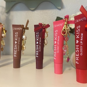 Puede incluir: Cinco tubos de bálsamo labial brillante Fresh Kiss de ColourPop en varios tonos, cada uno adornado con un charm. Los bálsamos labiales varían en color desde rosa claro hasta rojo intenso. Los charms incluyen cuentas, corazones y otros elementos decorativos.