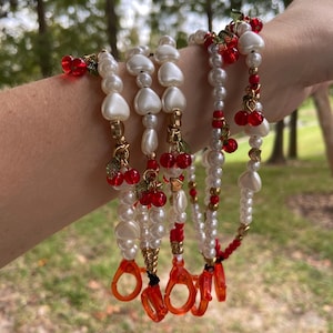 Peut inclure: Plusieurs cha&icirc;nes de lunettes perl&eacute;es avec des perles blanches, des perles en forme de c&oelig;ur et des accents de cerises rouges. Les cha&icirc;nes ont des accents m&eacute;talliques dor&eacute;s et des supports de lunettes orange. L'arri&egrave;re-plan est une sc&egrave;ne ext&eacute;rieure floue.