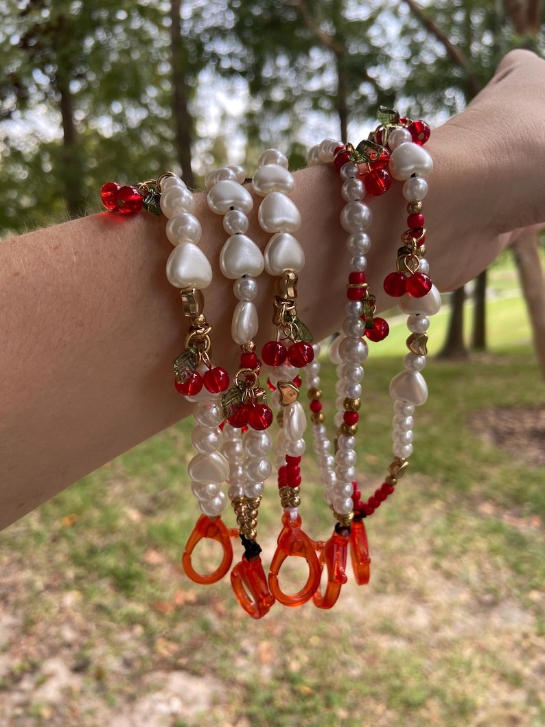 Peut inclure: Plusieurs cha&icirc;nes de lunettes perl&eacute;es avec des perles blanches, des perles en forme de c&oelig;ur et des accents de cerises rouges. Les cha&icirc;nes ont des accents m&eacute;talliques dor&eacute;s et des supports de lunettes orange. L'arri&egrave;re-plan est une sc&egrave;ne ext&eacute;rieure floue.