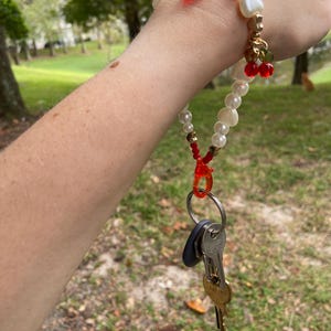 Peut inclure: Un bracelet porte-cl&eacute;s en perles et or avec des accents de cerises rouges. Le bracelet porte-cl&eacute;s contient un jeu de cl&eacute;s attach&eacute;es &agrave; un fermoir en plastique rouge. Le bracelet est port&eacute; au poignet.