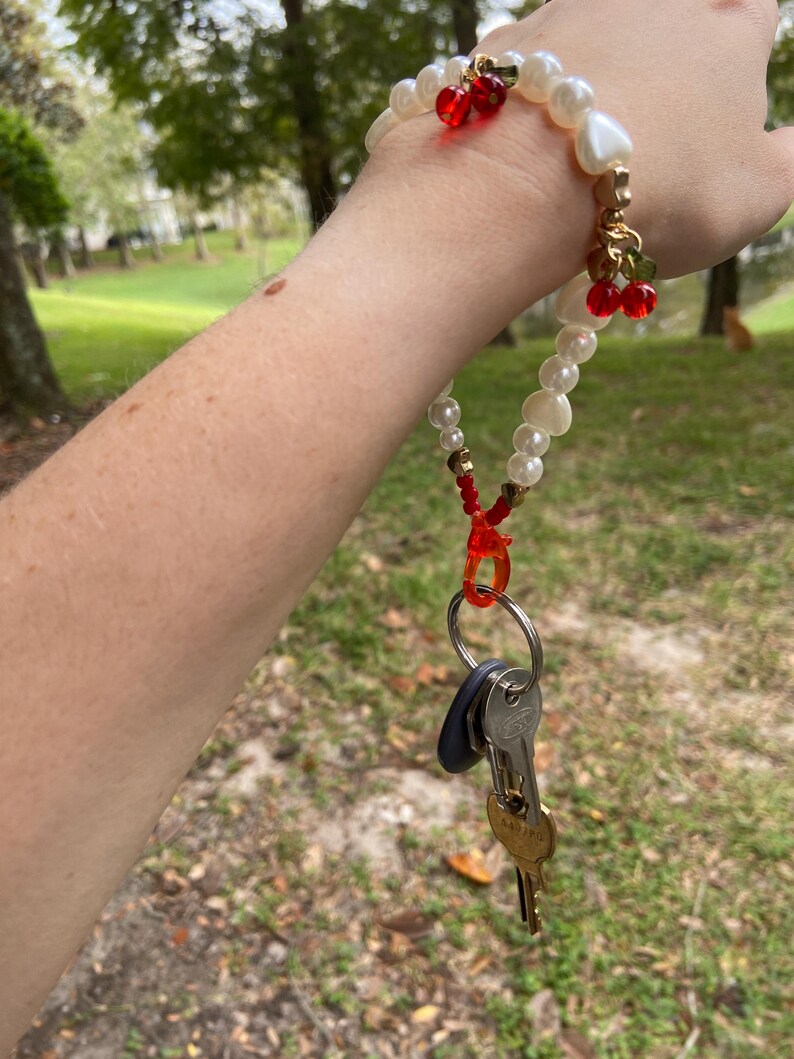 Peut inclure: Un bracelet porte-cl&eacute;s en perles et or avec des accents de cerises rouges. Le bracelet porte-cl&eacute;s contient un jeu de cl&eacute;s attach&eacute;es &agrave; un fermoir en plastique rouge. Le bracelet est port&eacute; au poignet.
