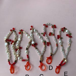 Peut inclure: Cinq lani&egrave;res perl&eacute;es avec des perles blanches, des perles en forme de c&oelig;ur, des accents rouges et des d&eacute;tails dor&eacute;s. Chaque lani&egrave;re a un fermoir orange vif. Les lani&egrave;res sont &eacute;tiquet&eacute;es A, B, C, D et E.